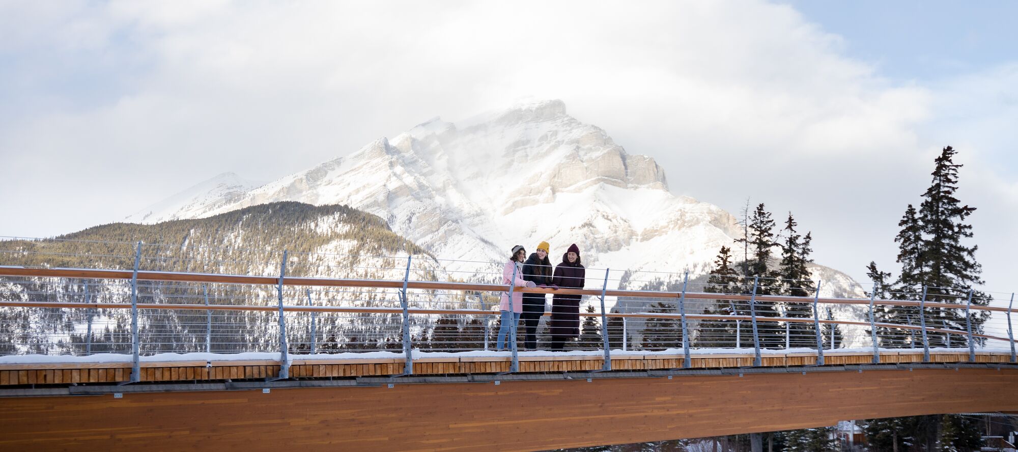 Three friends walk across the Nancy Pauw Bridge in winter with Cascade Mountain behind them and the Bow River below them in Banff National Park